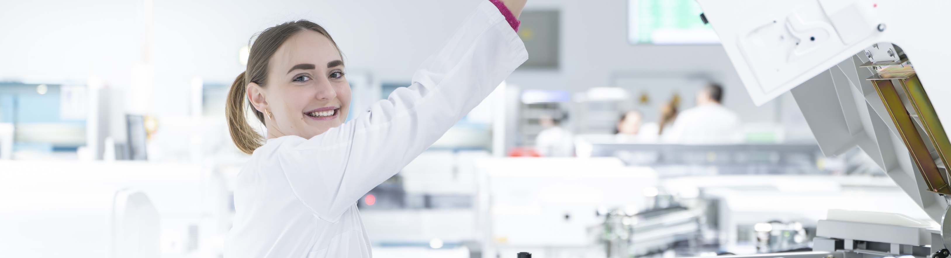 Eine Person in einem weißen Laborkittel hebt einen Arm in einem Laborumfeld und greift nach einem Analysengerät Die Frau lächelt. Im Hintergrund sind weitere Personen unscharf in Laborkitteln sowie Laborgeräte zu sehen.